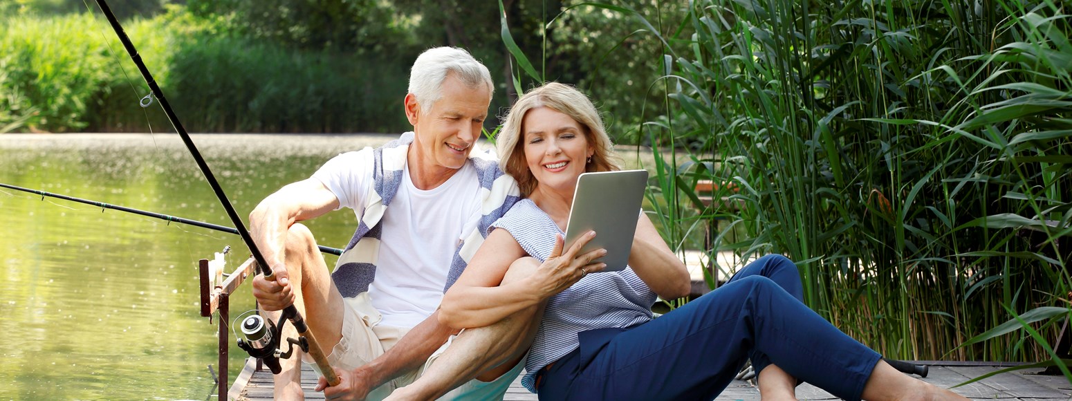 Älteres Paar sitzt zum Angeln auf einem Steg und betrachtet den Bildschirm eines Tablets (Geldanlagen)