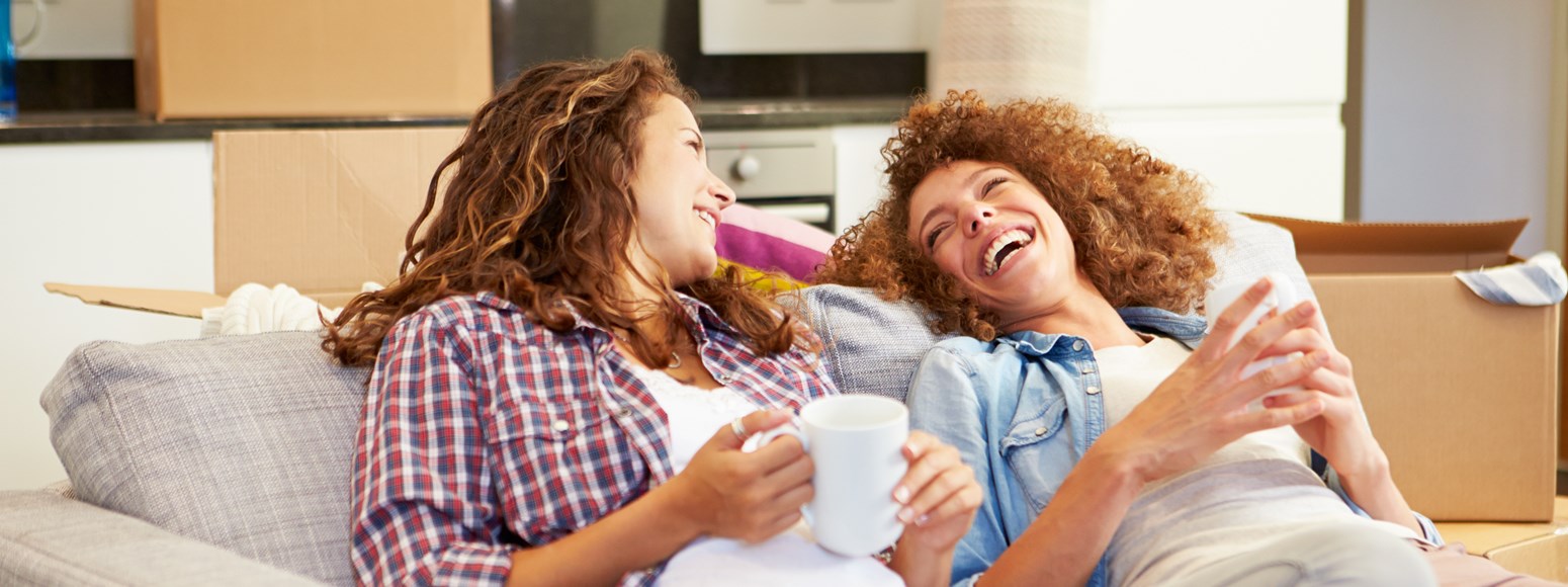 Zwei junge Frauen unterhalten sich lachend auf dem Sofa (Kunde wirbt Kunde)