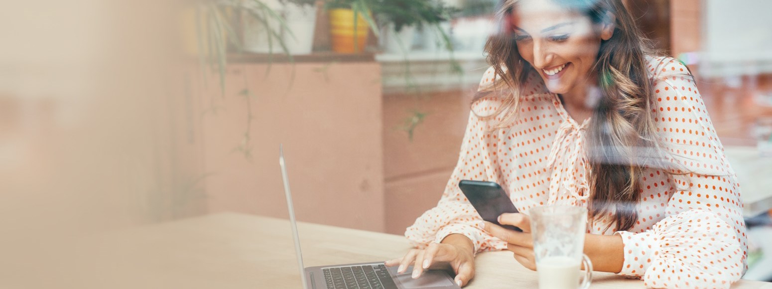 Junge Frau schaut lächelnd auf ein Smartphone und bedient einen Laptop (Digitaler Service) 